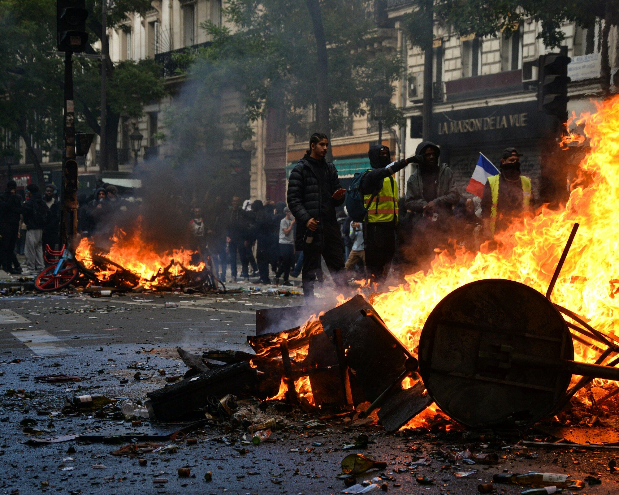 Brennende Barrikade aus Stühlen und Tischen bei Protesten in Paris, vermummte Demonstrierende mit Warnwesten und französischer Flagge im Rauch